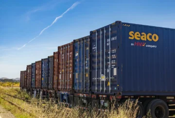 Row of vibrant cargo containers on train tracks under a clear blue sky.