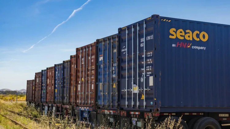 Row of vibrant cargo containers on train tracks under a clear blue sky.