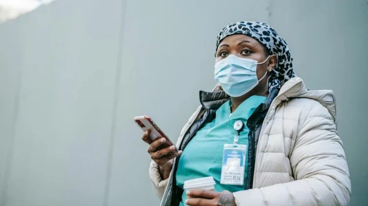 Serious adult African American nurse in warm clothes and protective face mask standing with cup of takeaway coffee and browsing mobile phone on street