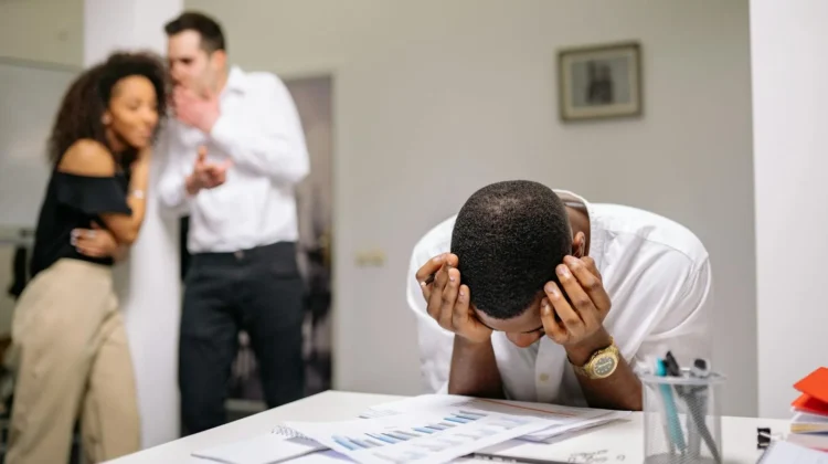 Despairing worker at office desk as colleagues gossip in background illustrating workplace bullying.
