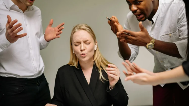 A stressed woman in an office surrounded by arguing coworkers highlighting workplace tension.